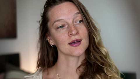 Video of a fair-skinned, middle-aged woman with long, wavy brown hair, wearing a white top and a gold necklace. She has a neutral expression, slightly smiling. The background is a blurred, neutral-toned room.