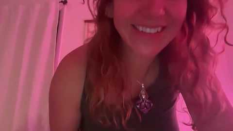 A video of a smiling, fair-skinned woman with curly brown hair, wearing a black tank top, taken in a bathroom with pink lighting.
