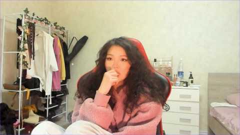 Video of an Asian woman with long, wavy black hair, wearing a pink sweater, sitting on a bed, gazing pensively. Background includes a white dresser, a clothes rack with colorful garments, and a gaming chair.