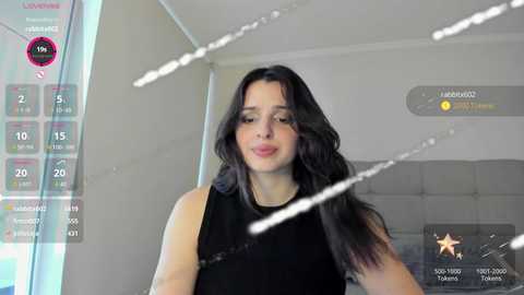 Video of a woman with long black hair, wearing a black sleeveless top, standing in a modern, minimalistic room with white walls and a gray couch.