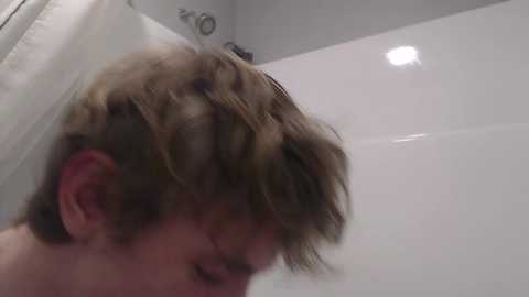 Video of a young man with tousled brown hair, smiling, in a white-tiled bathroom shower.