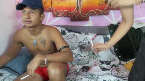 Video of a shirtless, tattooed young man with a dark complexion and red shorts, holding a controller, sitting on a bed with a black and white patterned comforter. Another person, partially visible, stands beside him.