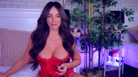 Video of a busty, curvaceous woman with long, wavy brown hair, wearing a red lace lingerie set, sitting on a bed with a brick wall background.