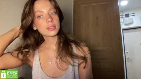 Video of a young woman with long brown hair, wearing a gray tank top, standing indoors by a wooden door. She has fair skin and a relaxed expression.