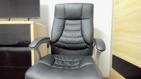 Video of a modern, black leather executive office chair with padded backrest and armrests, placed in a minimalist office setting with light wood paneling and a flat-screen TV on a beige cabinet.
