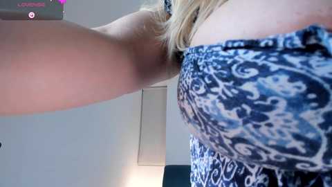 Video of a woman with fair skin and blonde hair, wearing a blue and white patterned strapless dress. Her arm is raised, showcasing her side profile. Background features a white wall and a lamp.