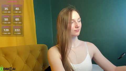 Video of a fair-skinned woman with long brown hair, wearing a white spaghetti-strap top, sitting on a green sofa. Background features a yellow-tufted chair and a digital display showing health metrics.