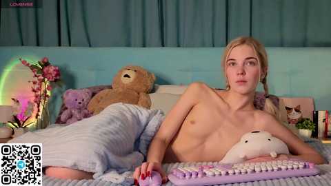 A video of a nude, fair-skinned, blonde woman lying on a bed with stuffed animals, wearing a light blue blanket, and holding a laptop.