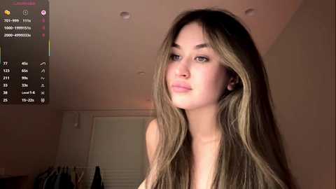 Video of a young woman with long, straight, light brown hair, light skin, and a slender build, looking slightly to the side. She is in a dimly lit bedroom with a TV displaying a streaming app interface in the background.