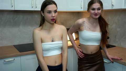 Video of two young women with fair skin, one with long brown hair, the other with long red hair, both wearing strapless white tops and high-waisted skirts, standing in a modern kitchen with white cabinets and a beige tiled backsplash.