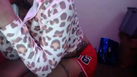 Video of a woman with light skin wearing leopard print top and red shorts, sitting on a bed with a tattoo on her arm, in a dimly lit room with a blue trash can and nightstand.