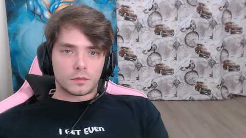 Video of a young man with fair skin, brown hair, and blue eyes, wearing a black shirt with \"I GET EVEN\" text and pink headphones, seated in front of a blue and brown patterned wall.