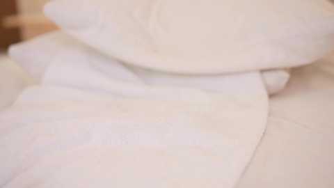 A close-up video of soft, white pillows stacked on a bed, with a blurred, neutral-colored background. The image focuses on the texture and arrangement of the pillows, creating a serene, cozy atmosphere.