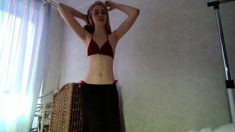 Video of a slender, light-skinned woman with long, straight hair, wearing a red bikini top and black sarong, stretching in a dimly lit room with a wicker chair and window with sheer curtains.