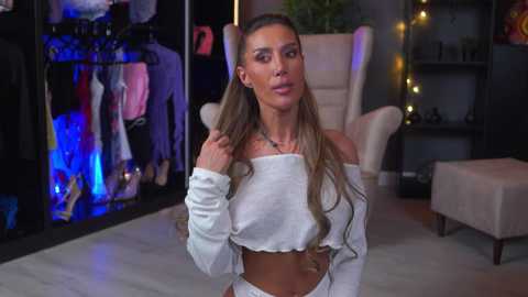 Video of a curvy, light-skinned woman with long, wavy blonde hair, wearing a white off-shoulder crop top, standing in a modern, dimly lit room with a white armchair, shelves, and clothes hanging in the background.