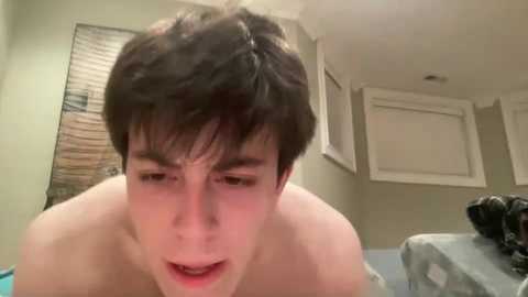 A video of a young man with short, dark hair, shirtless, looking slightly distressed, in a beige-walled room with a window and a small table.