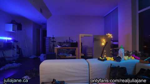 Video of a dimly lit, cluttered room with purple lighting, featuring a white couch with yellow flowers, a wooden table, and a fireplace with candles.