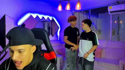 Video of a lively indoor scene featuring three young Asian men, one in a black cap and jacket, and two in casual attire, in a modern, brightly lit room with shark-themed decor and hanging lights.