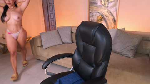 Video of a nude woman with small breasts and light skin, wearing only a white thong, standing in a living room. She is adjusting her hair. In the foreground, a black leather office chair with blue fabric.