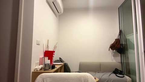 A video of a small, sparsely furnished hotel room with white walls, a beige sofa, and a red plastic chair. The room includes a wooden desk, a brown handbag hanging from the door, and an air conditioning unit on the wall.