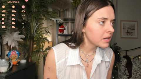 Video of a young woman with straight brown hair, wearing a white blouse, looking contemplative. Background features a cozy room with greenery, a bookshelf, and framed art.