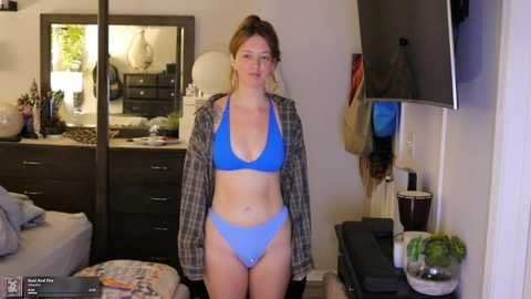 Video of a young woman with light skin and auburn hair, wearing a blue bikini and an open plaid shirt, standing in a modern bedroom with a large mirror, dresser, and flat-screen TV.