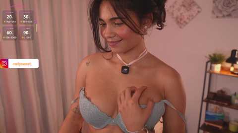 A video of a smiling Latina woman with medium skin tone, medium build, and dark hair, wearing a light blue bra and a pearl necklace, in a bedroom with pink curtains and a black shelf.