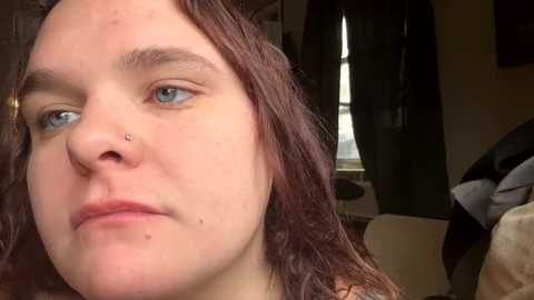 Video of a young Caucasian woman with fair skin, long red hair, and blue eyes. She wears a nose piercing and has acne on her face. The background shows a dimly lit room with dark curtains and a window.