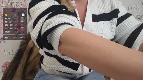 Video of a woman with light skin, wearing a white and black striped sweater, sitting with her arm raised, revealing a tattoo on her forearm. Background shows a blurry, pink and white wall with \"LIVE\" text.