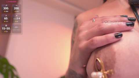 Video of a light-skinned woman with a tattooed hand on her breast, wearing black nail polish. Background features a blurred indoor setting with warm lighting.