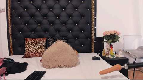 Video of a luxurious bedroom with a black, tufted headboard, leopard print pillow, beige fur pillow, a bouquet of pink roses, and a small toy giraffe on a white bed.