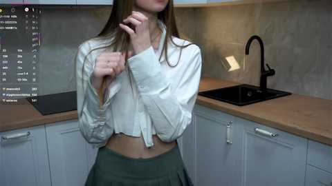 A video of a slender, light-skinned woman with long brown hair, wearing a white button-up shirt and green skirt, standing in a modern kitchen with light wood countertops and sleek black appliances.