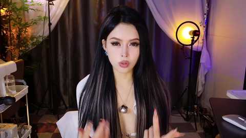 Video of a young Asian woman with long, straight black hair, wearing a white top, sitting in a dimly lit room with purple and white draped curtains, a spotlight, and a plant.