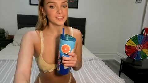 Video of a young woman with light skin and brown hair, wearing a beige bra and beige panties, holding a blue and orange soda can, sitting on a bed in a minimalistic bedroom.