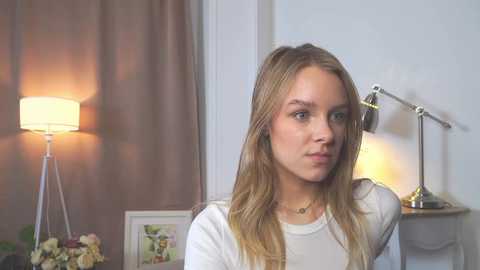 Video of a blonde woman with fair skin, wearing a white top, standing in a softly lit room with a lamp, flowers, and a shelf with a lamp and a framed photo.