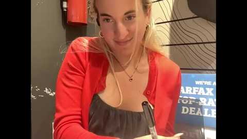 Media: A video of a blonde woman with a fair complexion, wearing a red cardigan over a black top, smiling while holding a microphone. Background features a \"We Are Not Fox News\" sign and a black wall with a red fire extinguisher.