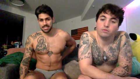 Video of two shirtless men with tattoos, one in gray briefs, sitting on a bed in a dimly lit bedroom.