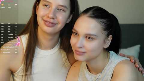 Video of two young women with light skin, straight hair, and wearing casual white tank tops, smiling closely. Background includes a blurred, indoor setting with a dark headboard and a digital screen displaying text.