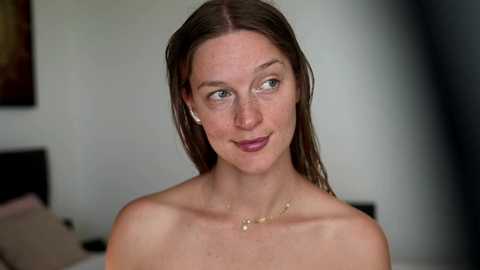 Video of a fair-skinned, slim woman with straight, shoulder-length brown hair, wearing minimal makeup and a delicate gold necklace. She has a calm expression and is seated in a softly lit room with a bed and blurred artwork in the background.
