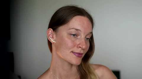 Video of a fair-skinned woman with long, straight brown hair, wearing a silver necklace, and silver earbuds, smiling softly with closed eyes, against a blurred, neutral background.