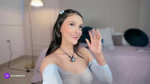 Video of a young woman with long black hair, light skin, and an off-shoulder light blue top, smiling and waving in a modern bedroom with soft lighting.