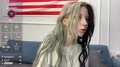A young woman with long, wet, blonde hair, wearing a grey t-shirt, sits on a blue couch in a dimly lit room. An American flag hangs behind her.
