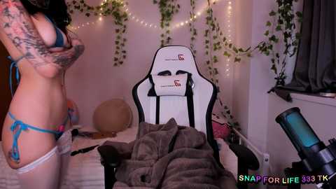 Video of a tattooed woman in blue lingerie and garter belt, standing in a cozy bedroom with fairy lights, a white gaming chair, and scattered clothes.
