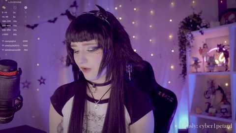 Video of a pale-skinned, androgynous individual with long black hair and blunt bangs, wearing a black choker and black dress. Background features a cozy room with fairy lights, a shelf with decorations, and a mic on the left.