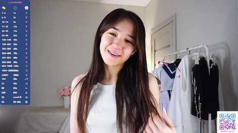 Video of a smiling Asian woman with long, straight black hair, wearing a white sleeveless top. Background features a white wall, a clothes rack with various garments, and a digital display screen with social media icons.