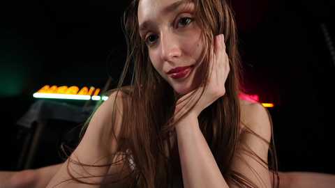 A video of a young woman with wet, long brown hair, fair skin, and light makeup, resting her chin on her hand. The background is dark with neon lights.