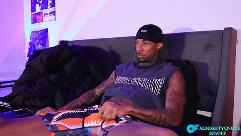 Video of a tattooed, muscular Black man with a shaved head, wearing a black cap and sleeveless shirt, lounging on a bed in a dimly lit room.
