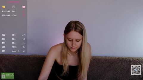 A video of a young woman with long blonde hair, wearing a black strapless dress, sitting on a sofa, looking down, in a dimly lit room.