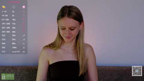 Video of a young Caucasian woman with long, straight blonde hair, wearing a black strapless dress, sitting on a grey couch. Background shows a digital screen displaying weather and time information.