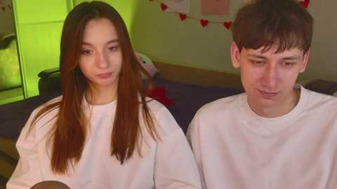 Video of a young Asian woman with long brown hair and a young Caucasian man with short brown hair, both wearing white T-shirts, sitting on a bed in a brightly lit room adorned with red heart decorations and stuffed animals.
